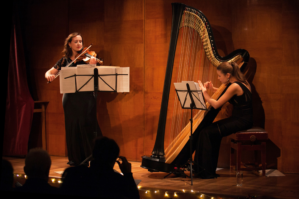 A violinist and harpist from the RCM perform on stage in the wood-paneled lecture theatre at Kensington Central Library